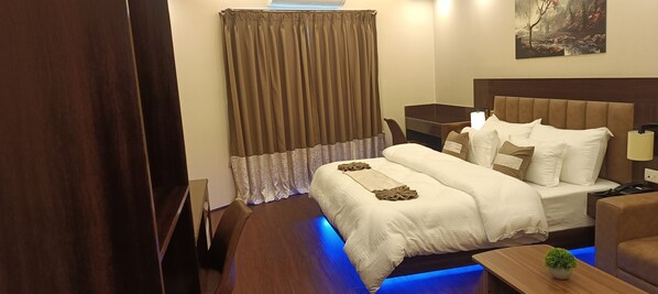 Een hotelkamer met een groot bed, een bureau, een stoel, een kleine tafel met een plant en een schilderij aan de muur.