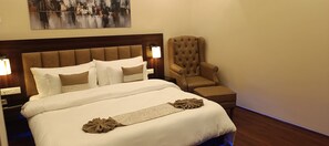 A neatly made bed with white and brown bedding, a brown upholstered armchair, and a wall-mounted lamp. - XCELSIOR HOTEL AND SPA BY ROH (Shillong)