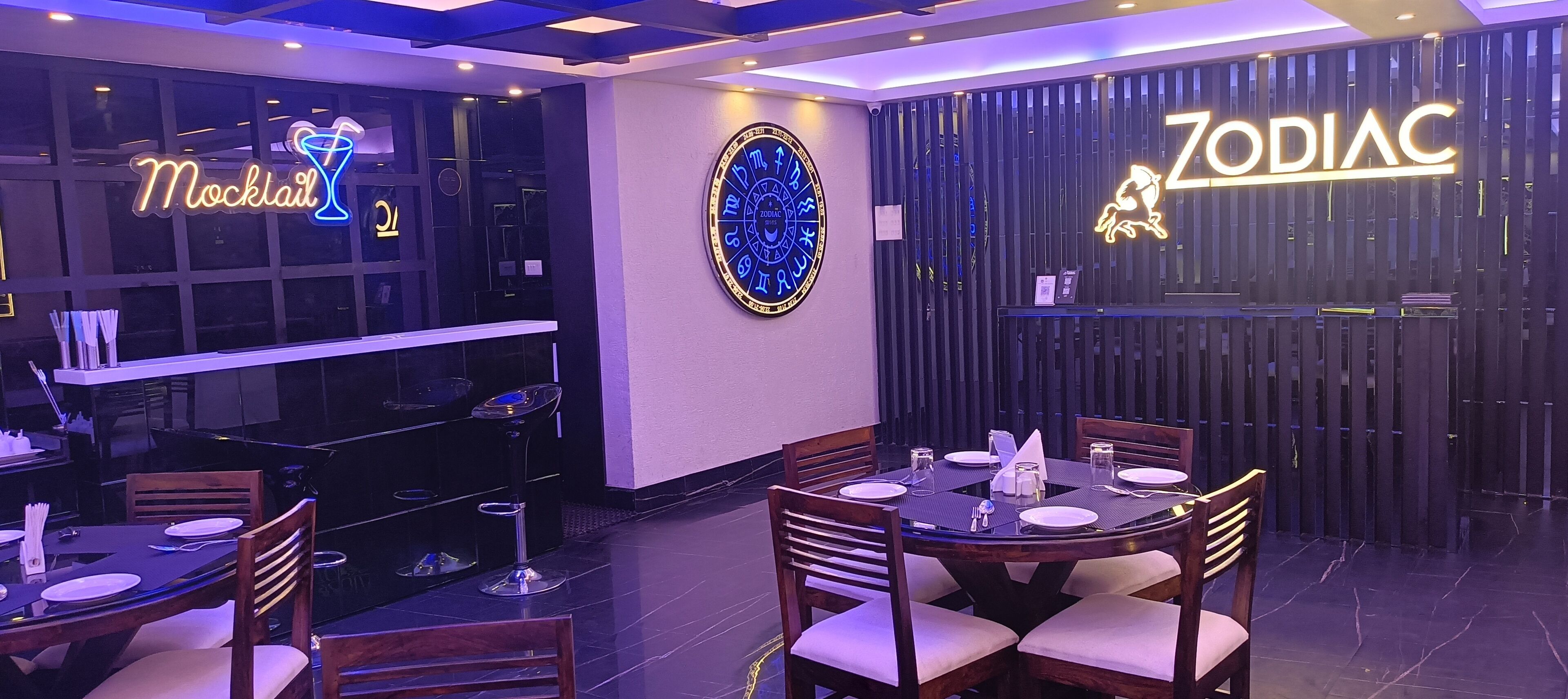 A restaurant with a neon sign reading "Mocktail" and "Zodiac". Tables are set with white tablecloths and silverware.