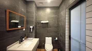 Un baño moderno con una pared de azulejos oscuros, suelo de madera, un lavabo blanco, un inodoro y un espejo con toallero.