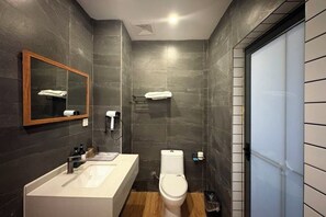 Un baño moderno con una pared de azulejos oscuros, suelo de madera, un lavabo blanco, un inodoro y un espejo con toallero.