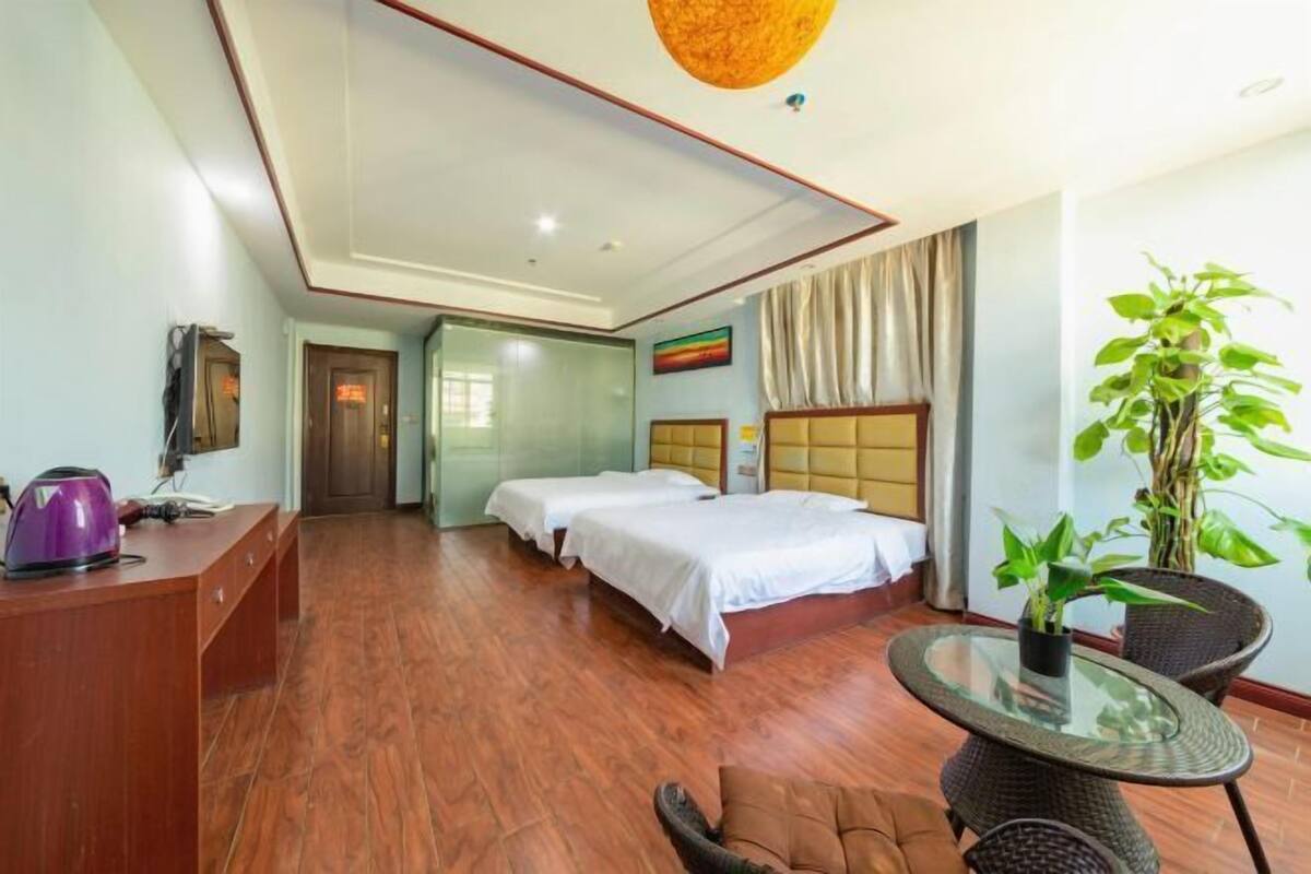 A hotel room with two beds, a desk with a kettle, a glass table, and a potted plant.