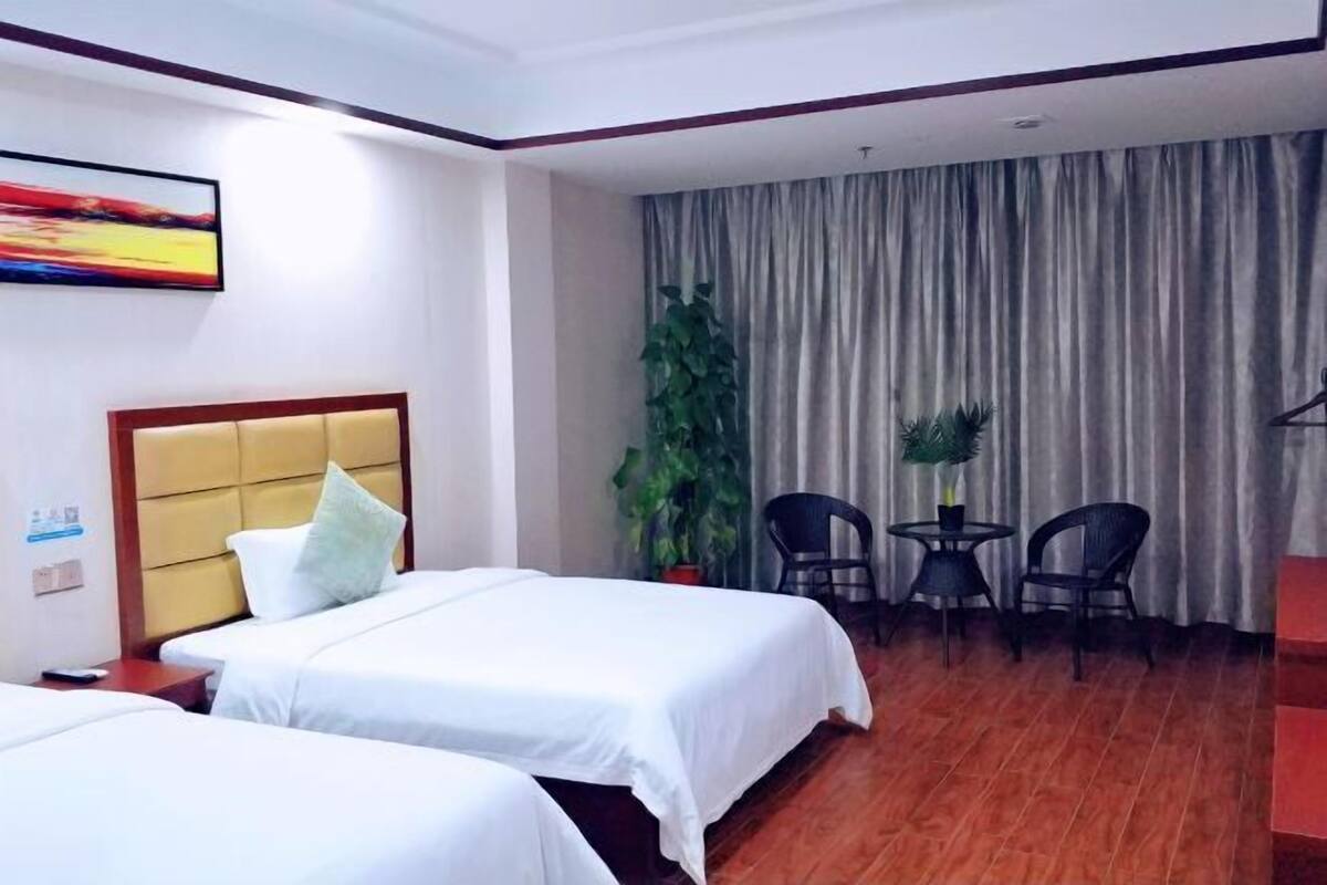 A hotel room with two beds, a large window with curtains, a plant, and a small table with chairs.