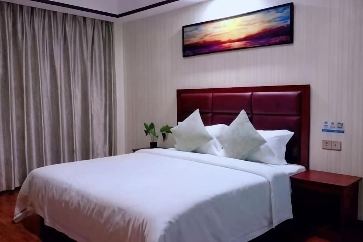 A hotel room with a large bed, a nightstand, a painting on the wall, and a window with curtains.