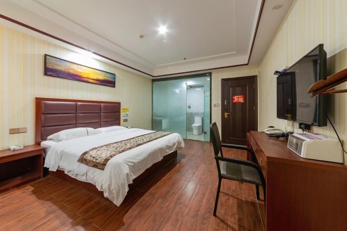 A hotel room with a bed, a desk, a chair, a television, and a bathroom visible through a glass door.