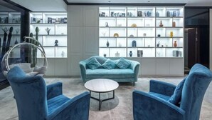 Un salon moderne avec un canapé turquoise, deux fauteuils bleus, un fauteuil suspendu en verre et une table basse blanche. Un grand meuble blanc avec différentes décorations se trouve à l’arrière-plan.
