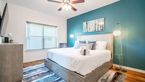 A modern bedroom with a large bed, a ceiling fan, a painting on the wall, and a bedside lamp. - Cowboys & Rangers Game-Day Townhome | Free Parking (Arlington)