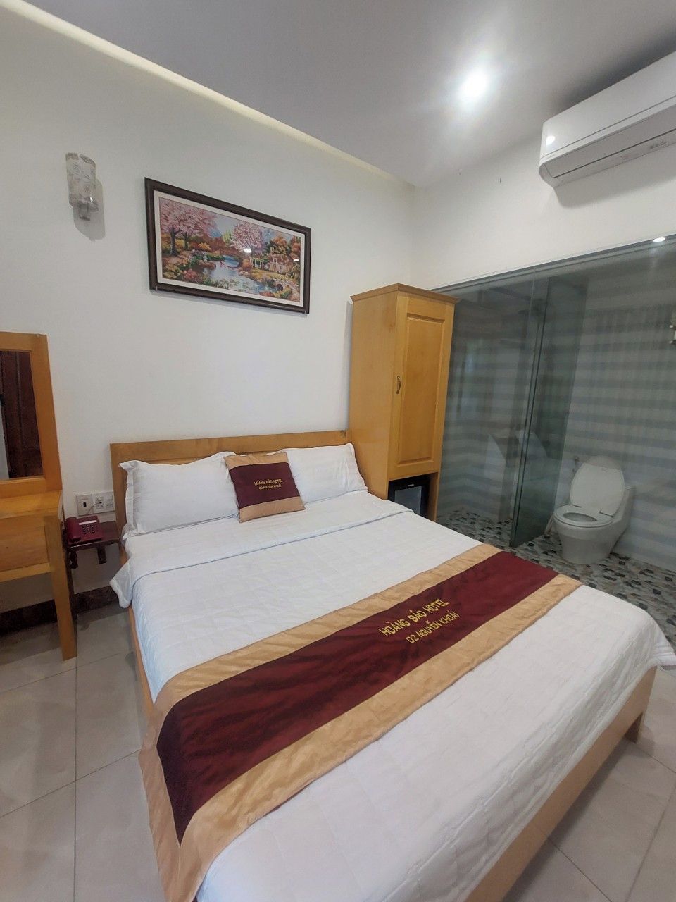 A hotel room with a bed, a wooden wardrobe, a bathroom with a glass shower enclosure, and a painting on the wall.