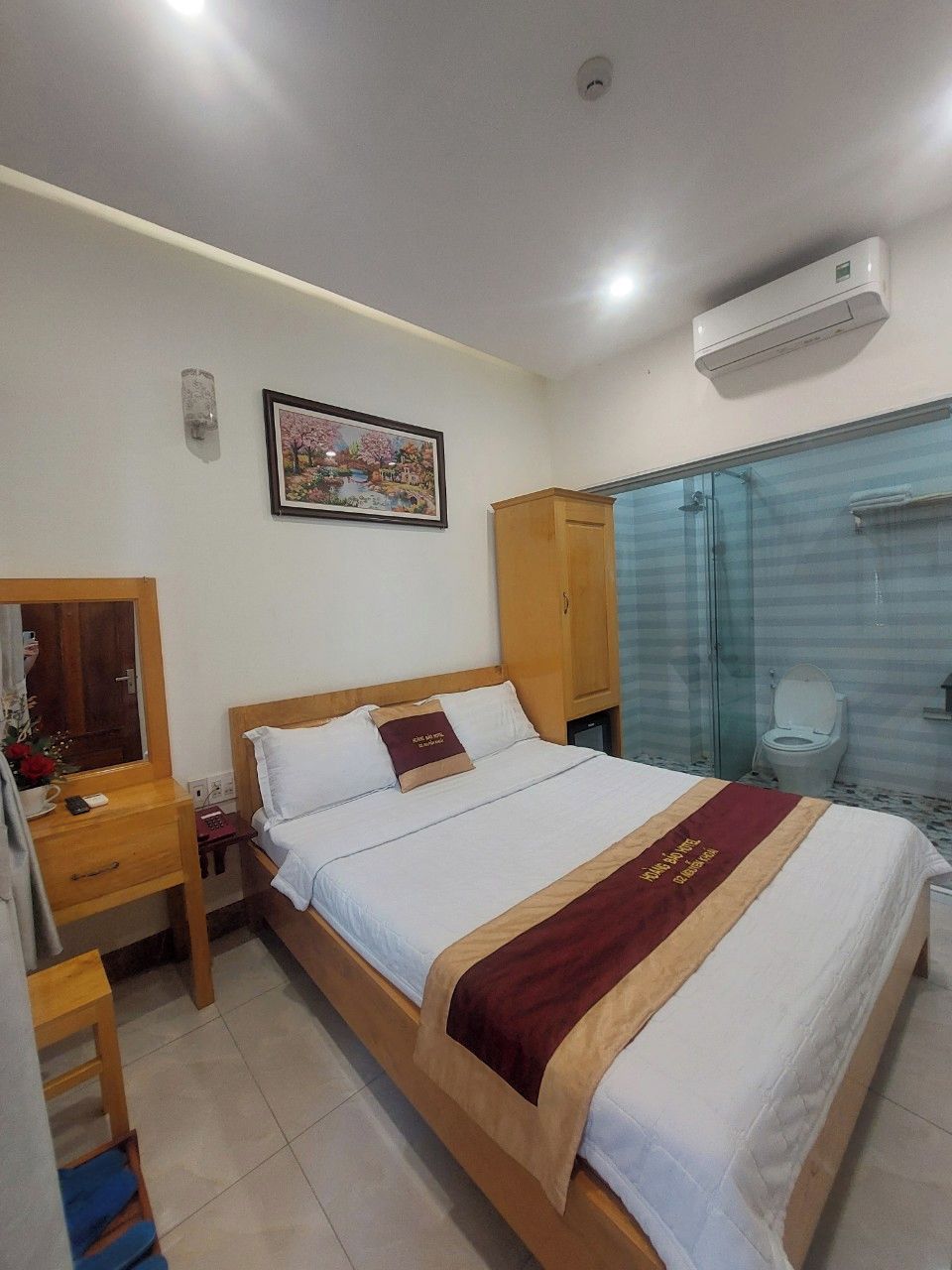 Habitación de hotel con cama, armario de madera, baño con cabina de ducha de cristal y aire acondicionado instalado en la pared.