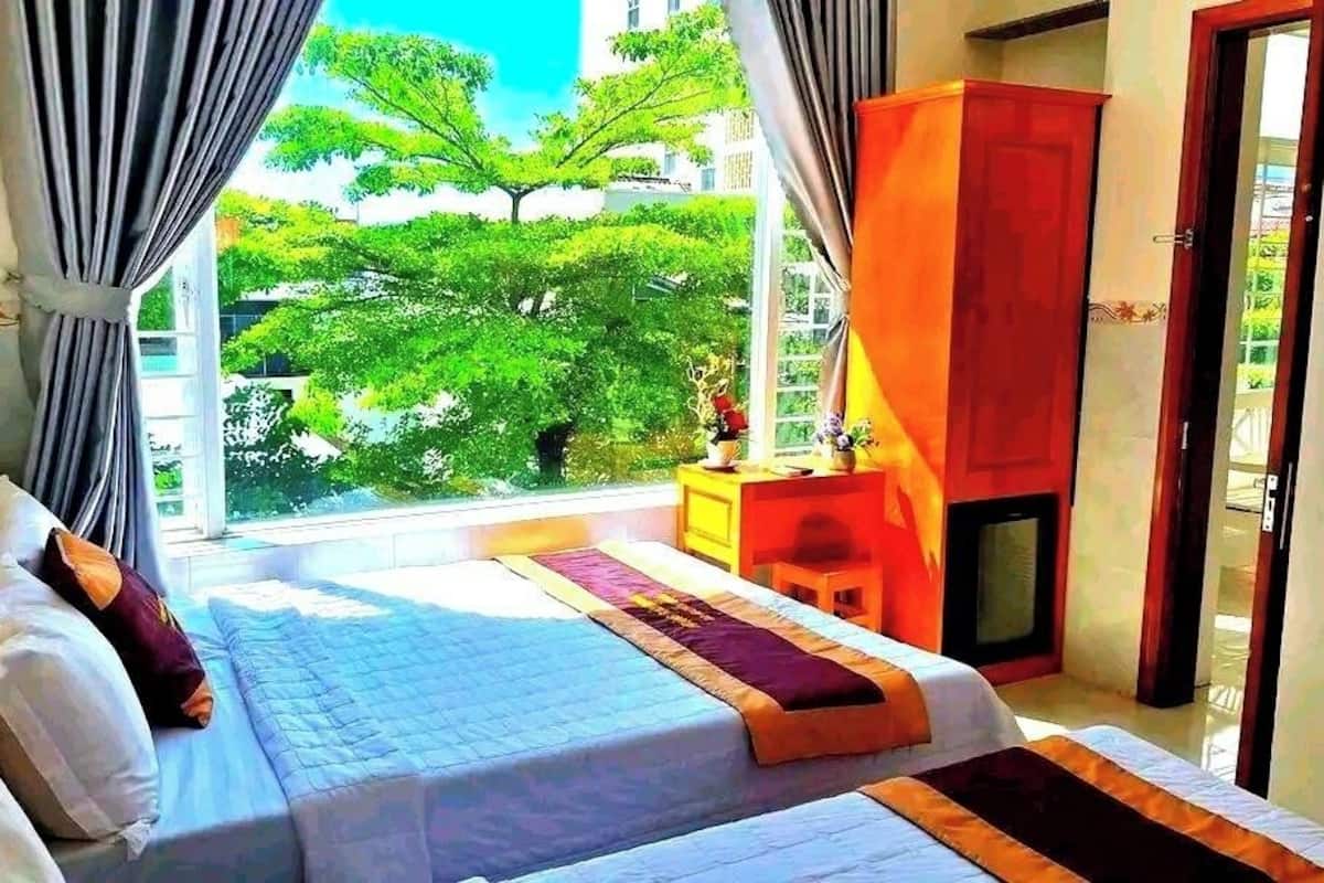 A hotel room with a bed, a small table with a lamp, and a view of greenery outside.
