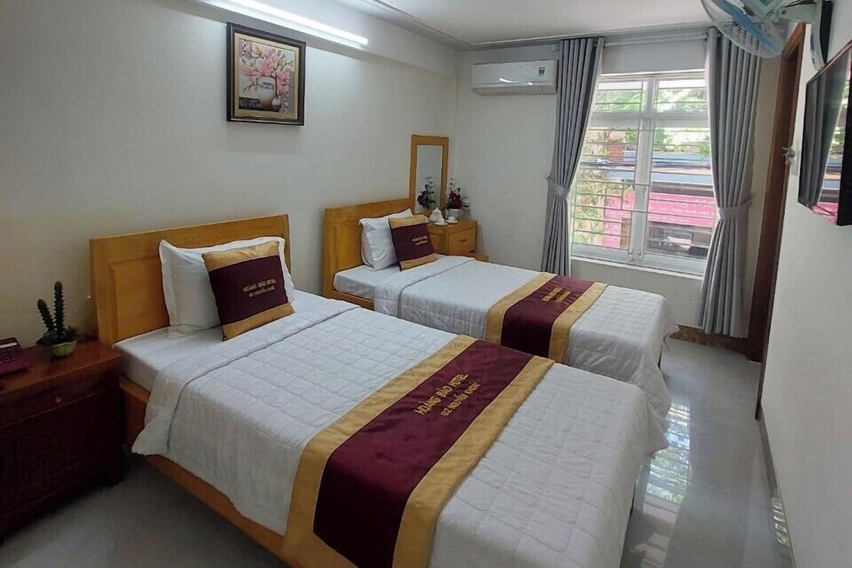 A hotel room with two beds, a bedside table, a nightstand, a wall-mounted air conditioner, a ceiling fan, a window with curtains, and a framed picture on the wall.