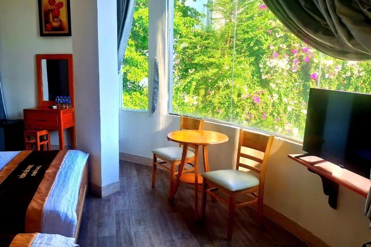 A hotel room with a bed, a desk with a chair, a TV, and a window with a view of trees and flowers.
