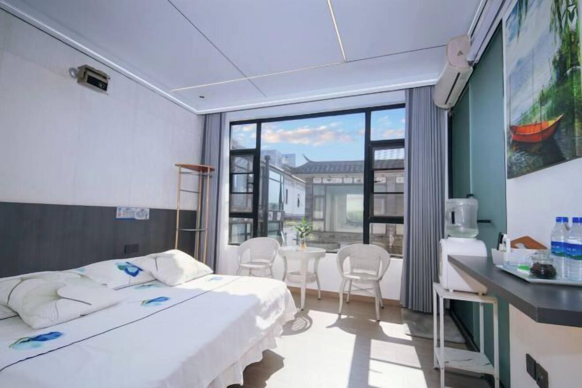 A hotel room with a bed, a desk with a chair, a small table, and a large window with a view of buildings.