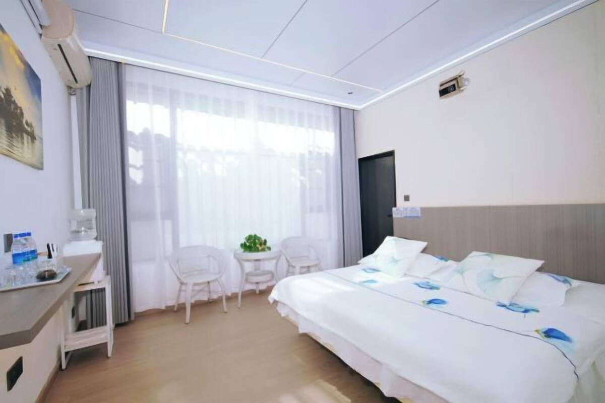 A hotel room with a bed, a desk with a chair, a small table with a plant, and a window with curtains.