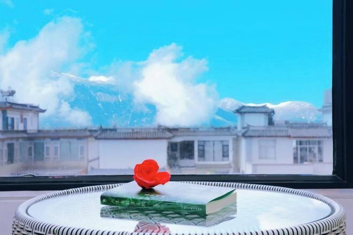 A book on a round table with a red flower, a window with a view of buildings, and a clear blue sky.
