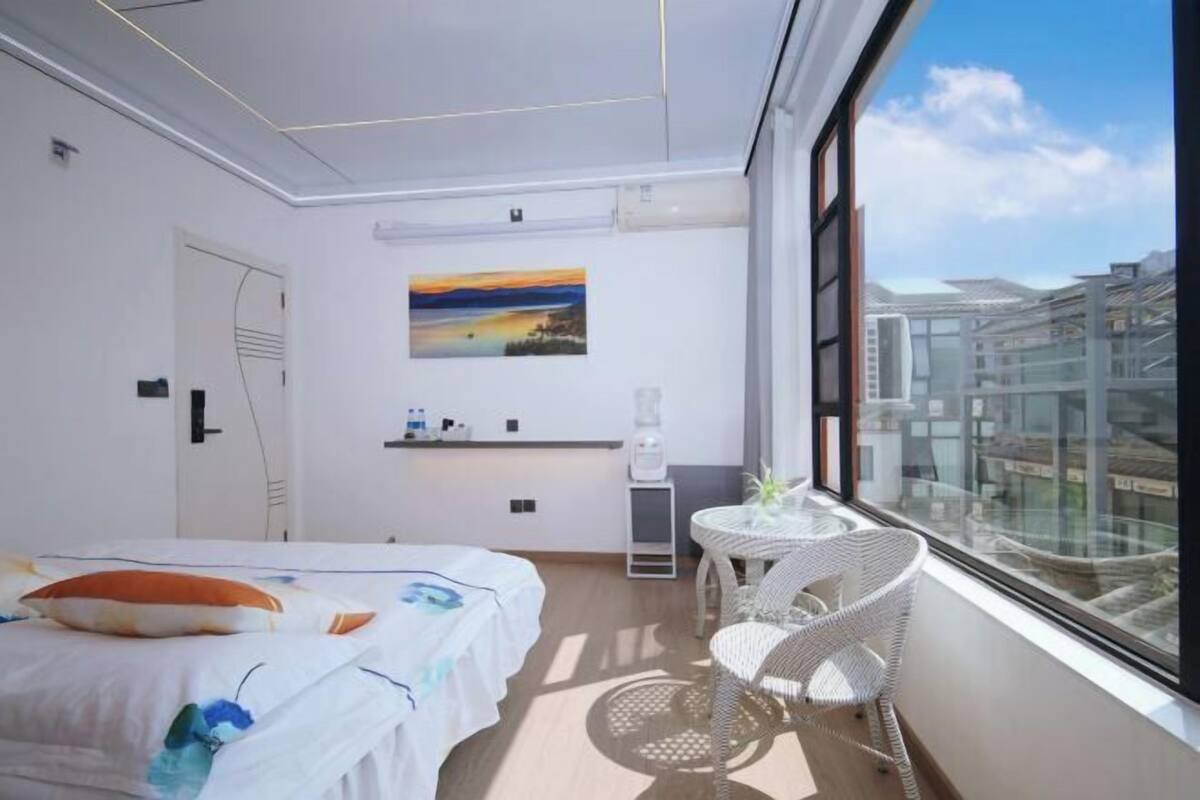 A modern hotel room with a bed, a small table, a chair, a painting on the wall, and a city view through the window.