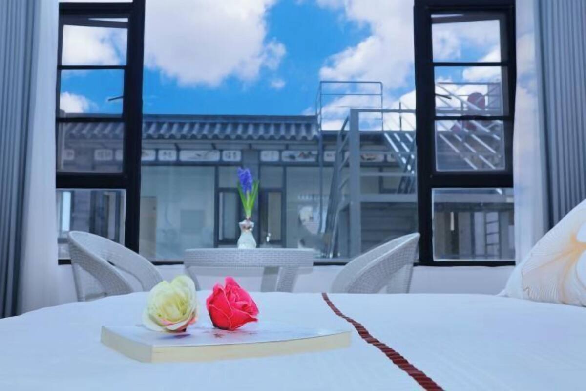 A hotel room with a bed, a table with a book and a flower, and a view of a building through large windows.