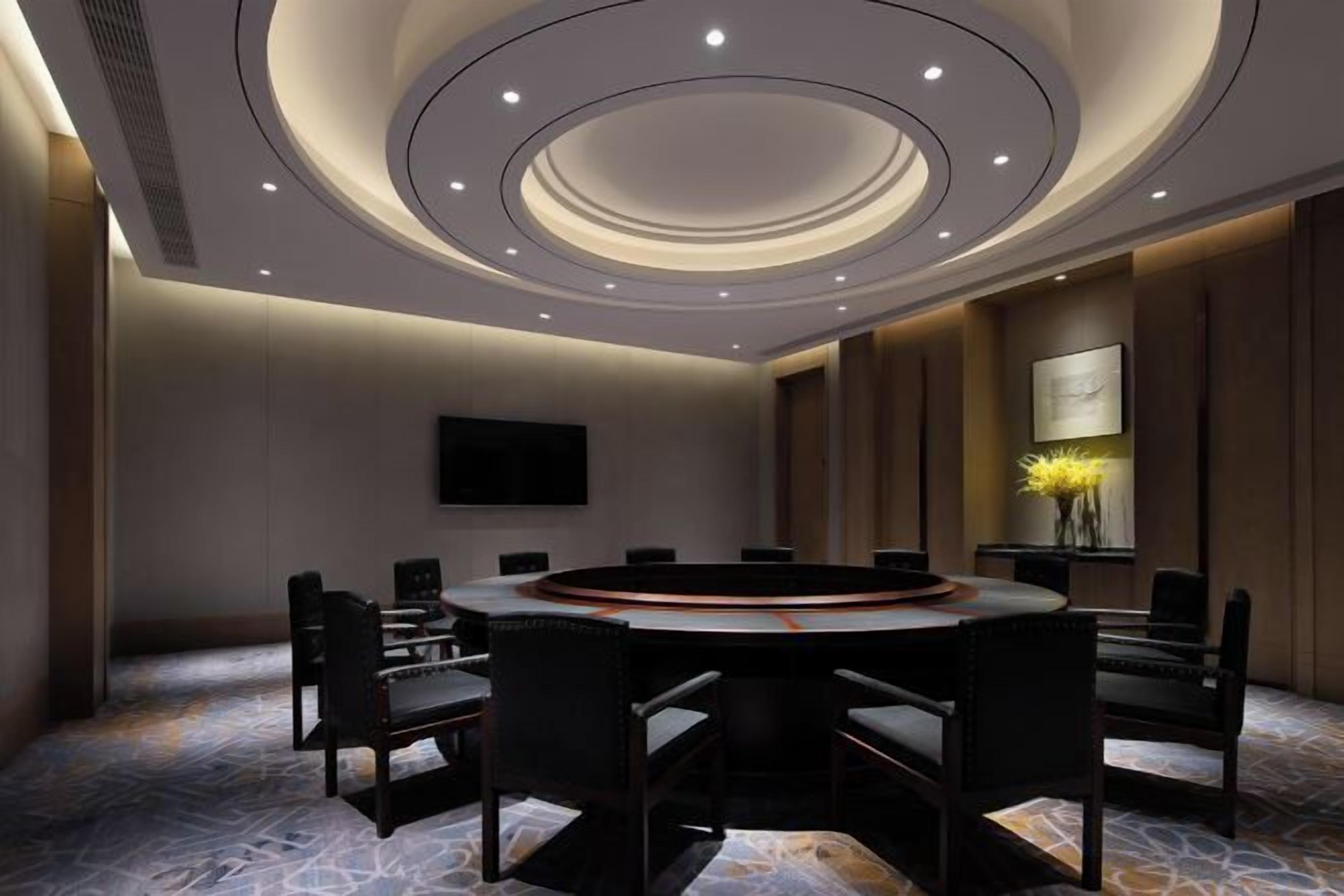 Una sala de conferencias con una mesa redonda, sillas, un televisor de pantalla plana y un jarrón decorativo con flores.