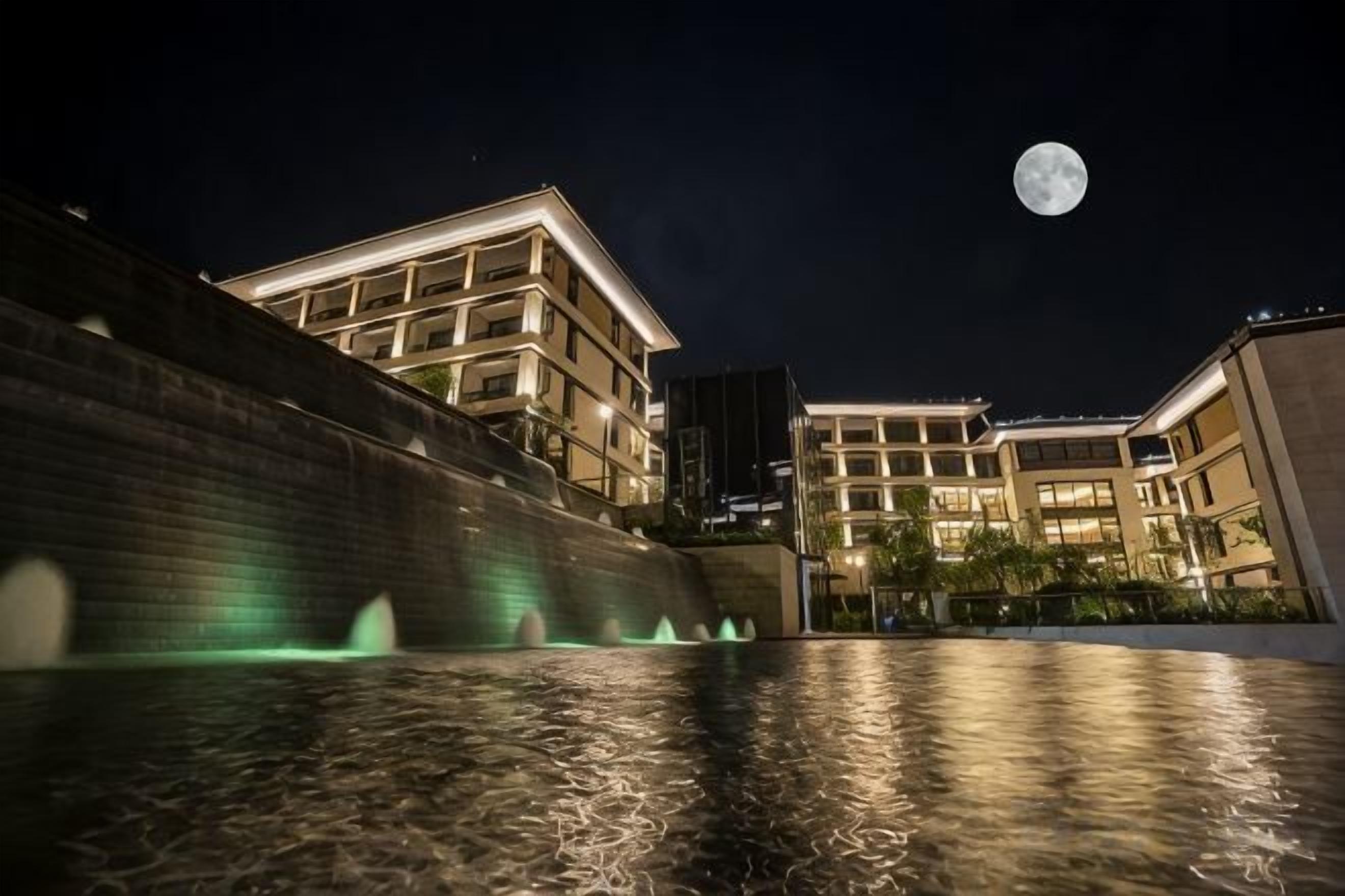 A nighttime view of a building with a water feature in front, a full moon in the sky, and illuminated windows.