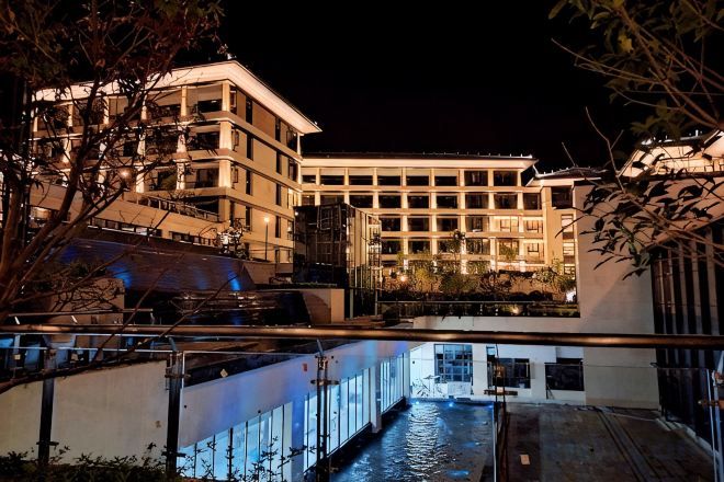 Un hotel moderno con fachada de vidrio, una fuente en primer plano y un exterior bien iluminado durante la noche.