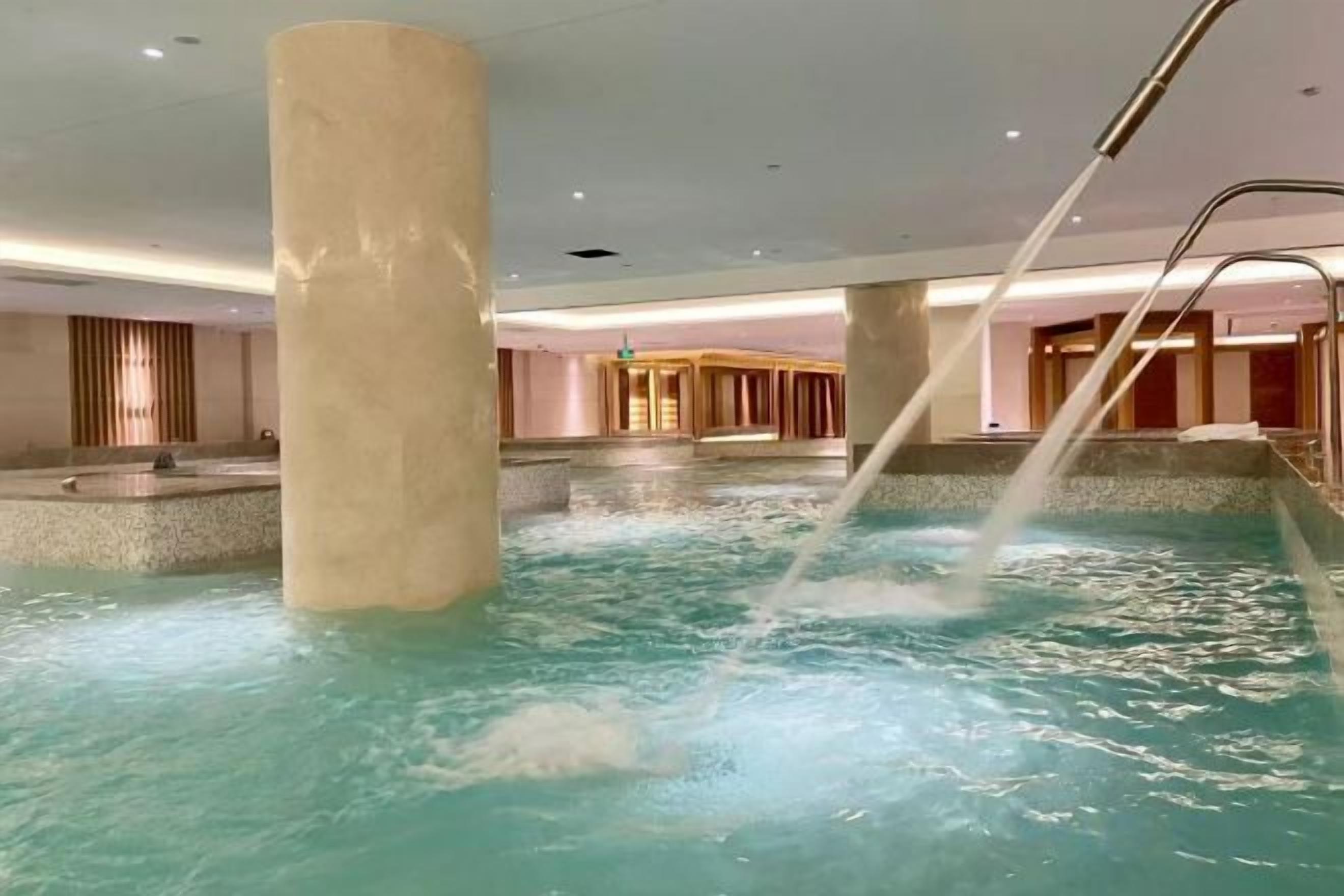 Una piscina interior con chorros de agua, un column y una zona de recepción al fondo.
