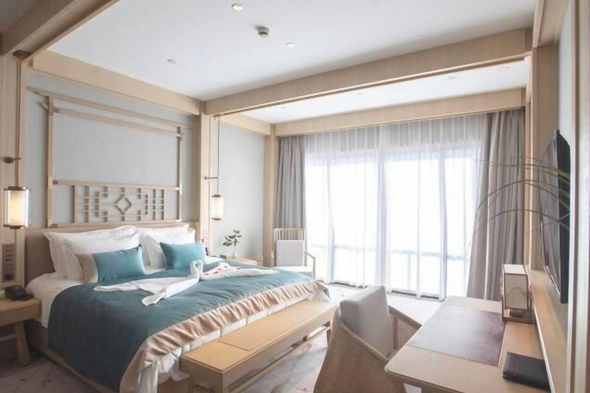 Een hotelkamer met een bed, nachtkastje, stoel, bureau en groot raam.