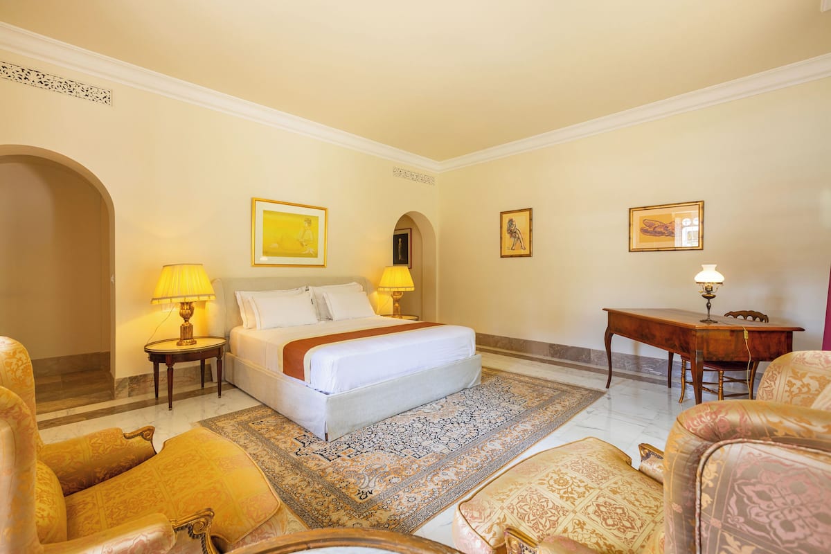 Habitación de hotel con una cama grande, dos mesitas de noche con lámparas, un escritorio de madera y una alfombra estampada.