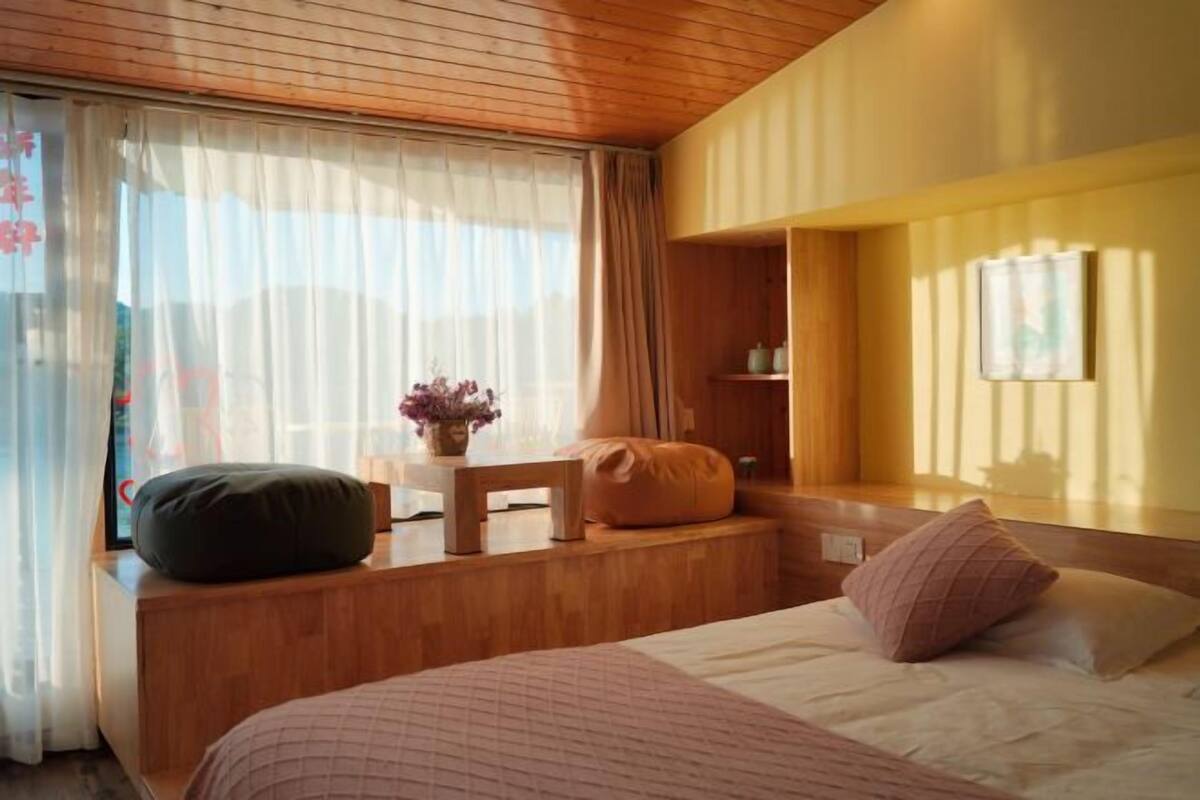 Una camera da letto con testiera in legno, un letto con copripiumino rosa e bianco, una panca di legno con cuscino, un tavolino con un vaso di fiori e una mensola con oggetti decorativi.