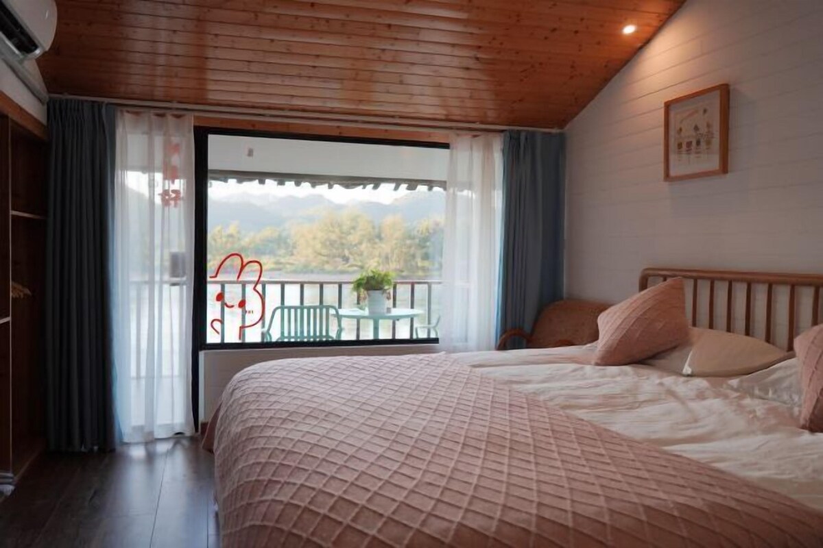 A bedroom with a large bed, a balcony with a table and chairs, and a view of trees and mountains.