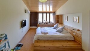 Een slaapkamer met een houten vloer, een bed met kussens, een televisie aan de muur en een groot raam met gordijnen.