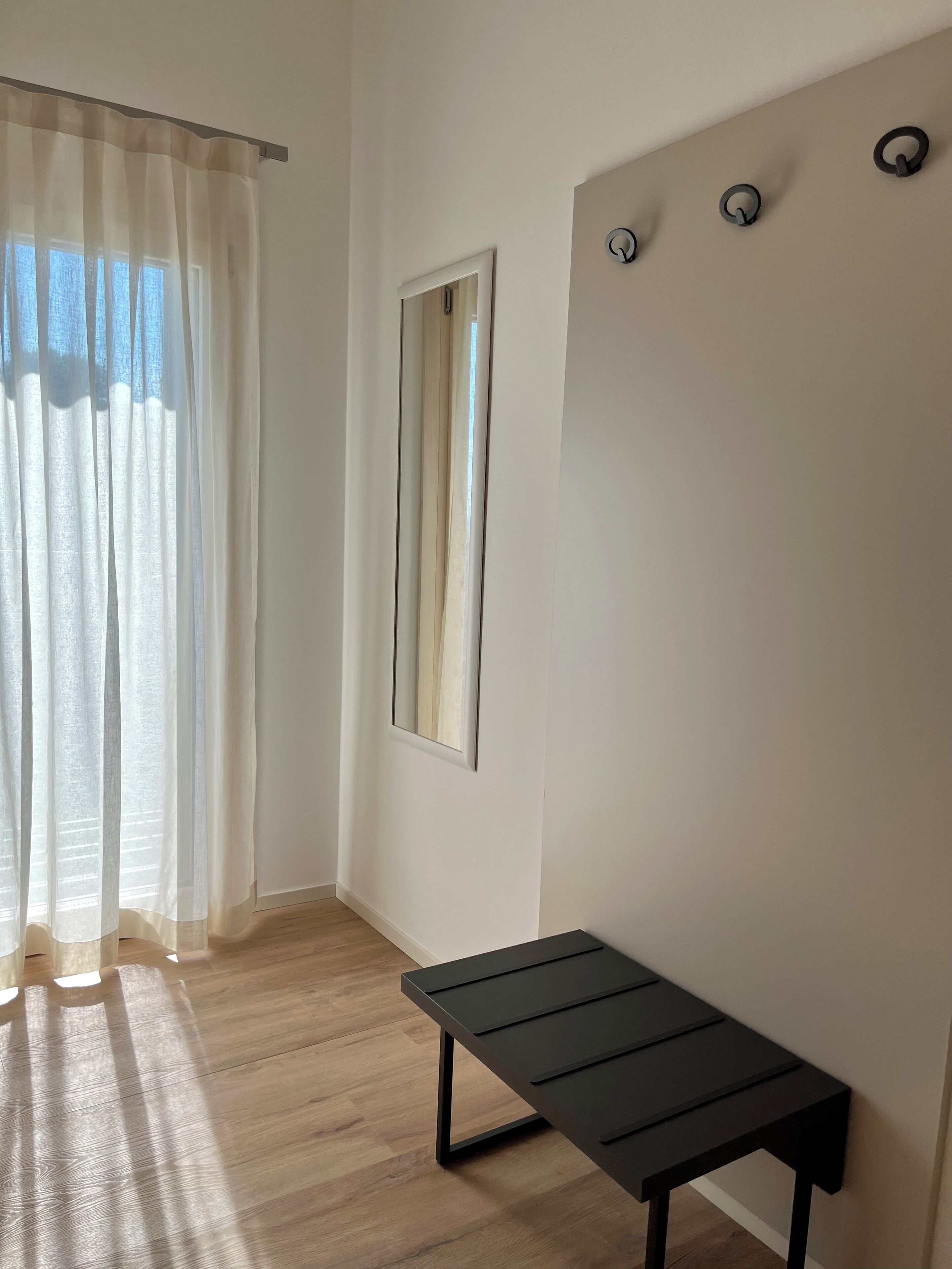 A room with a wooden floor, a black bench, a mirror, and a window with sheer curtains.