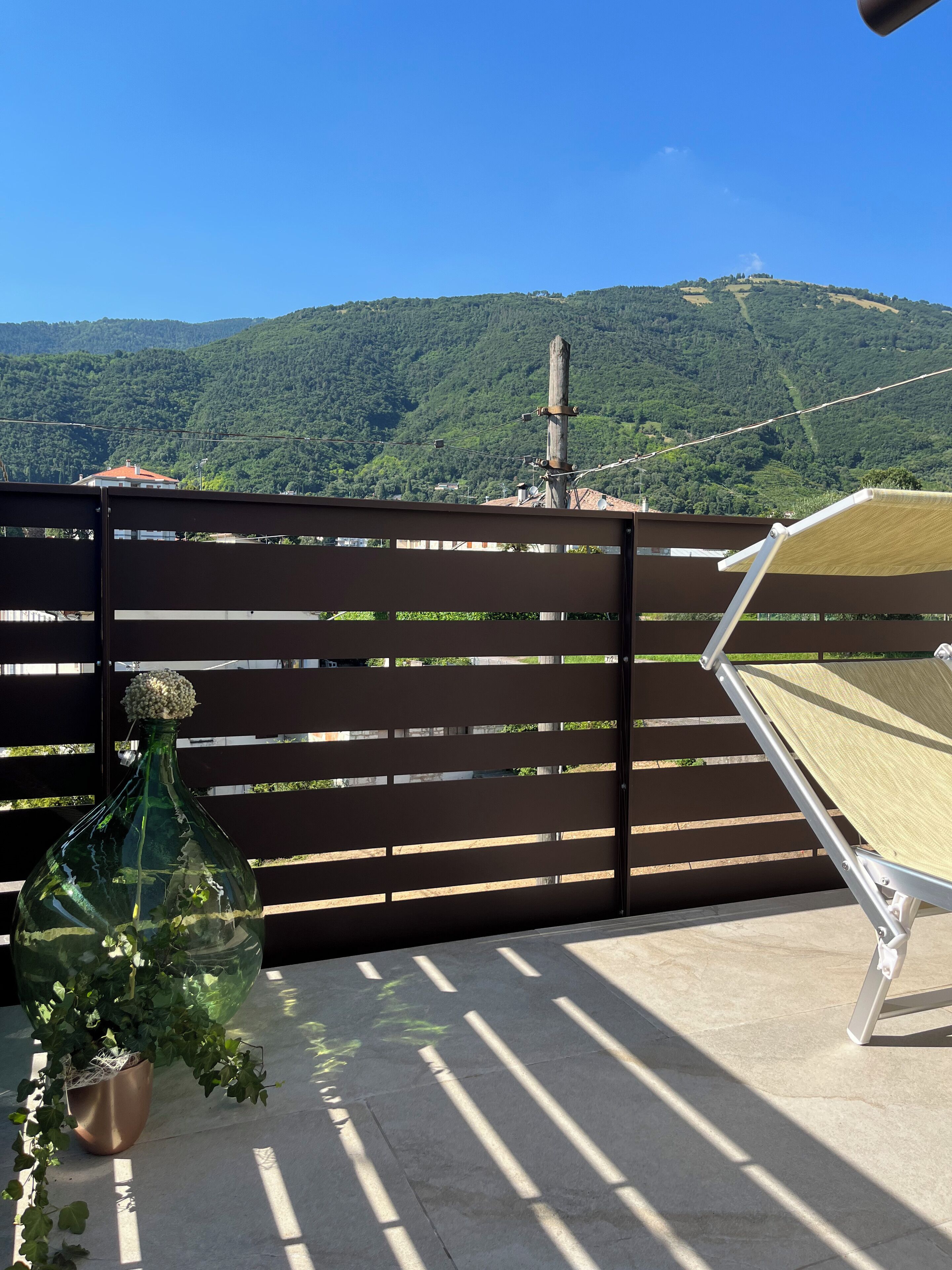 Un balcon donnant sur une colline verdoyante, avec une clôture en bois, une chaise en métal et un pot de fleurs.