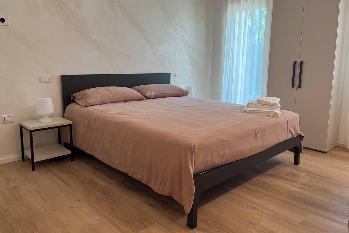 Une chambre à coucher avec un lit, une table de chevet, une lampe, une armoire et une fenêtre avec des rideaux.
