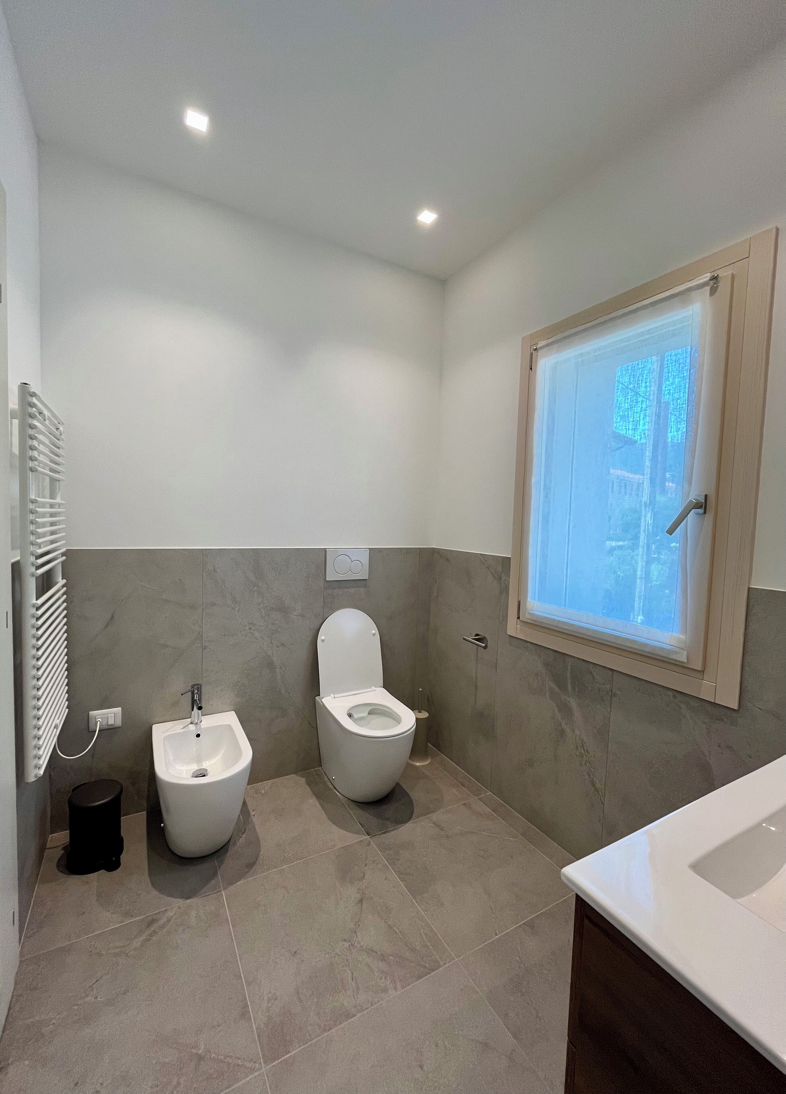 A modern bathroom with two toilets, a bidet, a window with a view, and a towel rack.