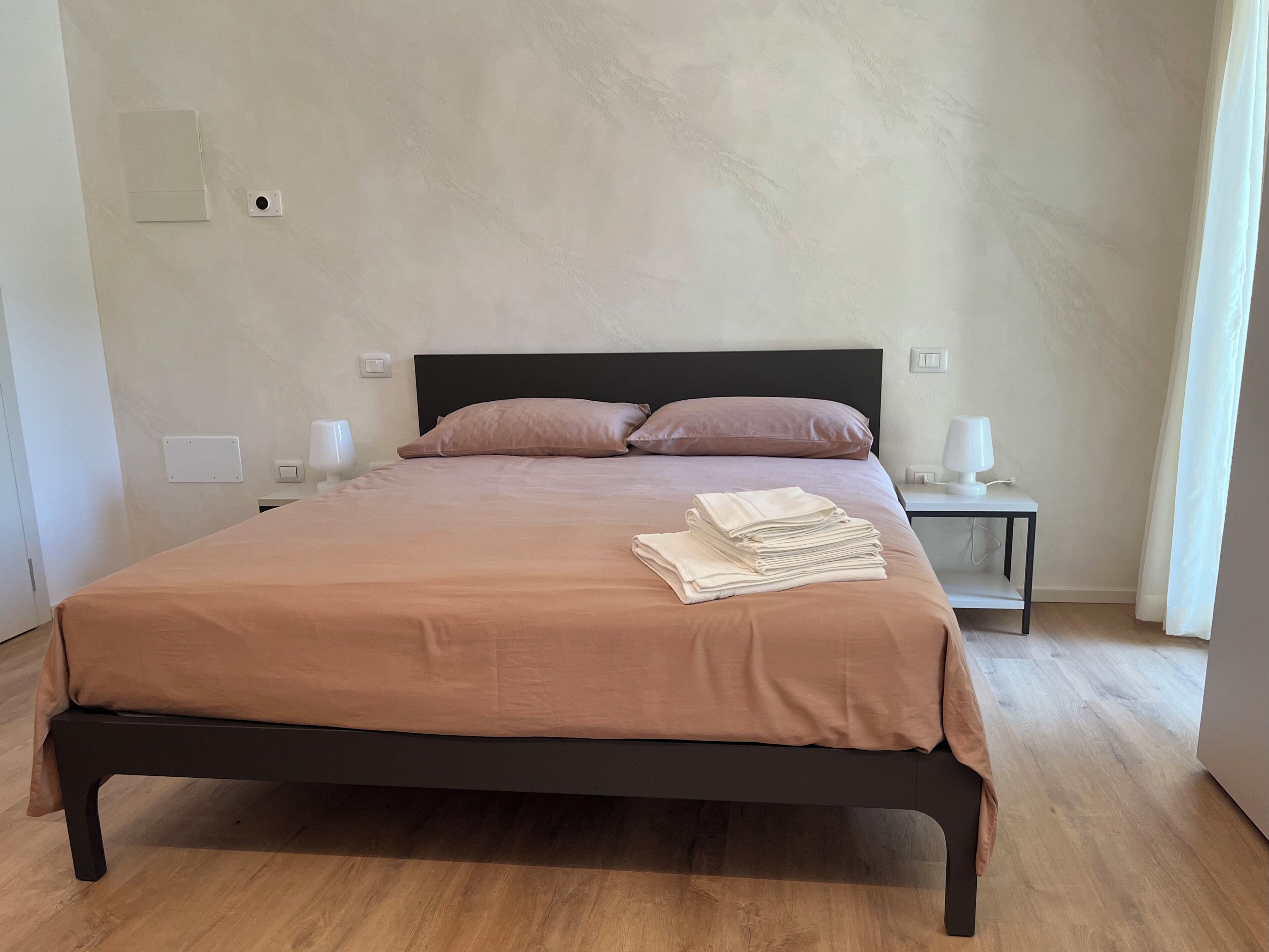 A neatly made bed with a brown bedspread and pillows, a small bedside table with a lamp, and a wall socket.