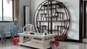 A modern living room with a glass-top coffee table, two chairs, a large wooden bookshelf, and a red trash bin. - Taining Luxiang Nanhui Hotel (Jinhu Prairie) (Taining)
