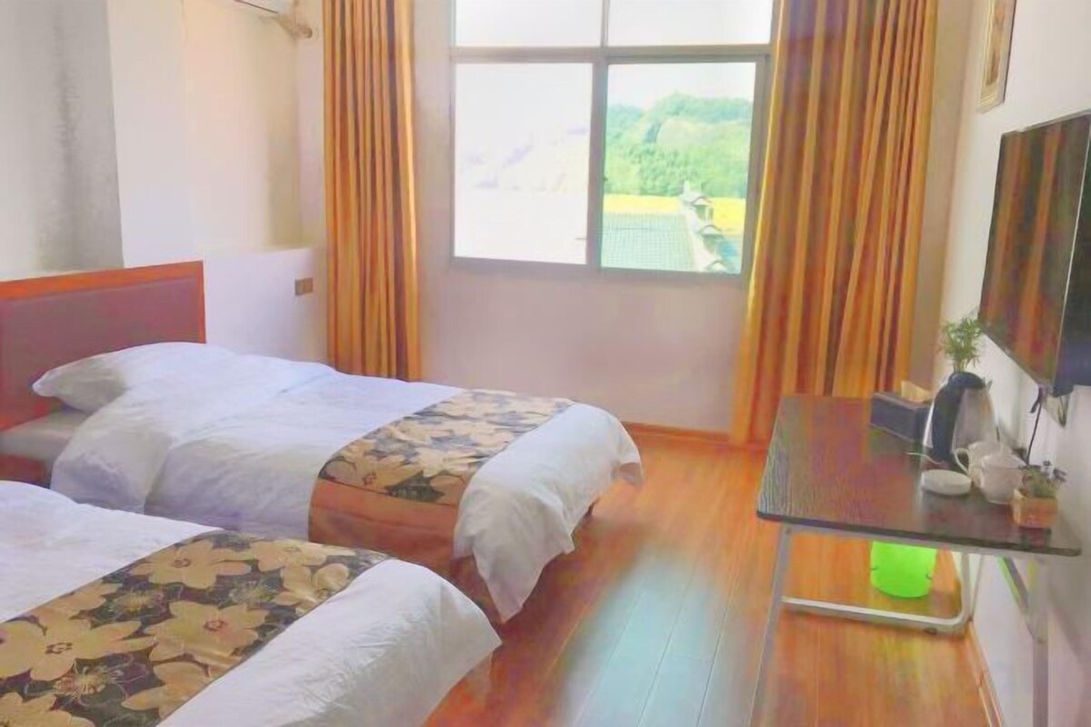 A room with two beds, a small table, a window with curtains, and a view of greenery outside.