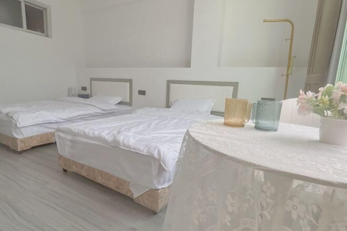 Una habitación de hotel con dos camas individuales, una mesa redonda con mantel de encaje, un florero con flores y una lámpara de pie dorada.