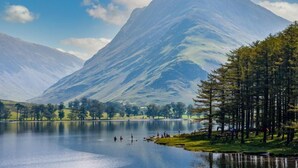 Un lago tranquilo con montañas al fondo, rodeado de árboles y una zona verde donde la gente disfruta de la vista.