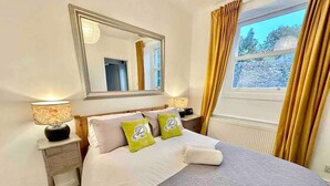 Un dormitorio ordenado con una cama grande, una mesita de noche con lámpara, un espejo sobre la cama y una ventana con cortinas.