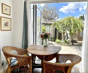 Una habitación con una mesa de madera, sillas de mimbre, un florero con flores y vistas a un jardín a través de un ventanal.