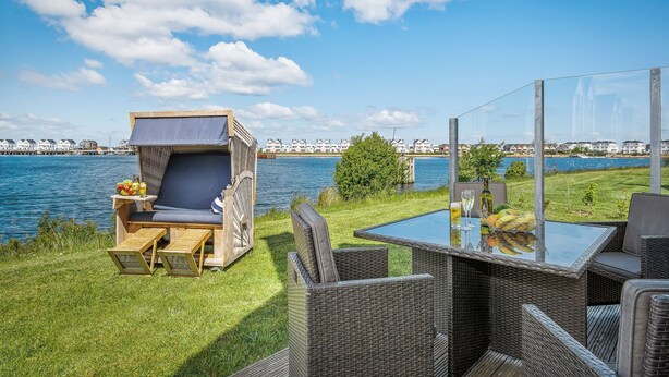 An outdoor seating area with wicker furniture, a glass table, and a view of a body of water and buildings in the background. - Hansekogge - detached, first row on the water, sauna, whirlpool tub, shower (Olpenitz)