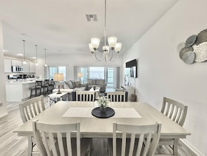 Une cuisine moderne avec des meubles blancs, un coin repas doté d’une table et de chaises en bois, et un salon avec un canapé et des fauteuils.