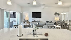 A modern kitchen with a central island, a dining area with wooden chairs, a ceiling fan, and a TV mounted on the wall. - Lost Shaker of Salt - 3/3 w/ 4-Seater Golf Cart in Moultrie Creek, The Villages! (The Villages)