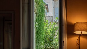 Hotelkamer met een bed, een stoel, een tafel en een open deur die leidt naar een buitenruimte met uitzicht op groen en een gebouw.