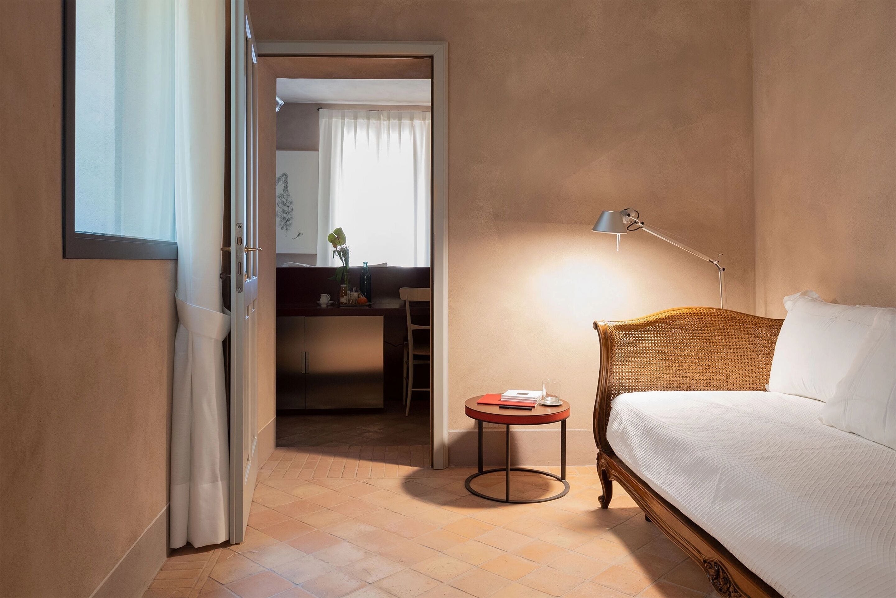 A hotel room with a bed, a bedside table, a lamp, a small round table, and a view of the hallway and another room.