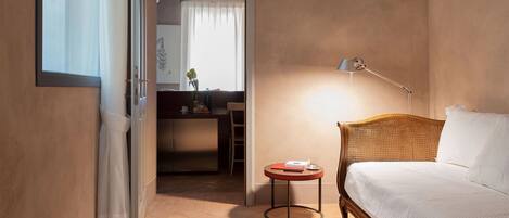 A hotel room with a bed, a bedside table, a lamp, a small round table, and a view of the hallway and another room.