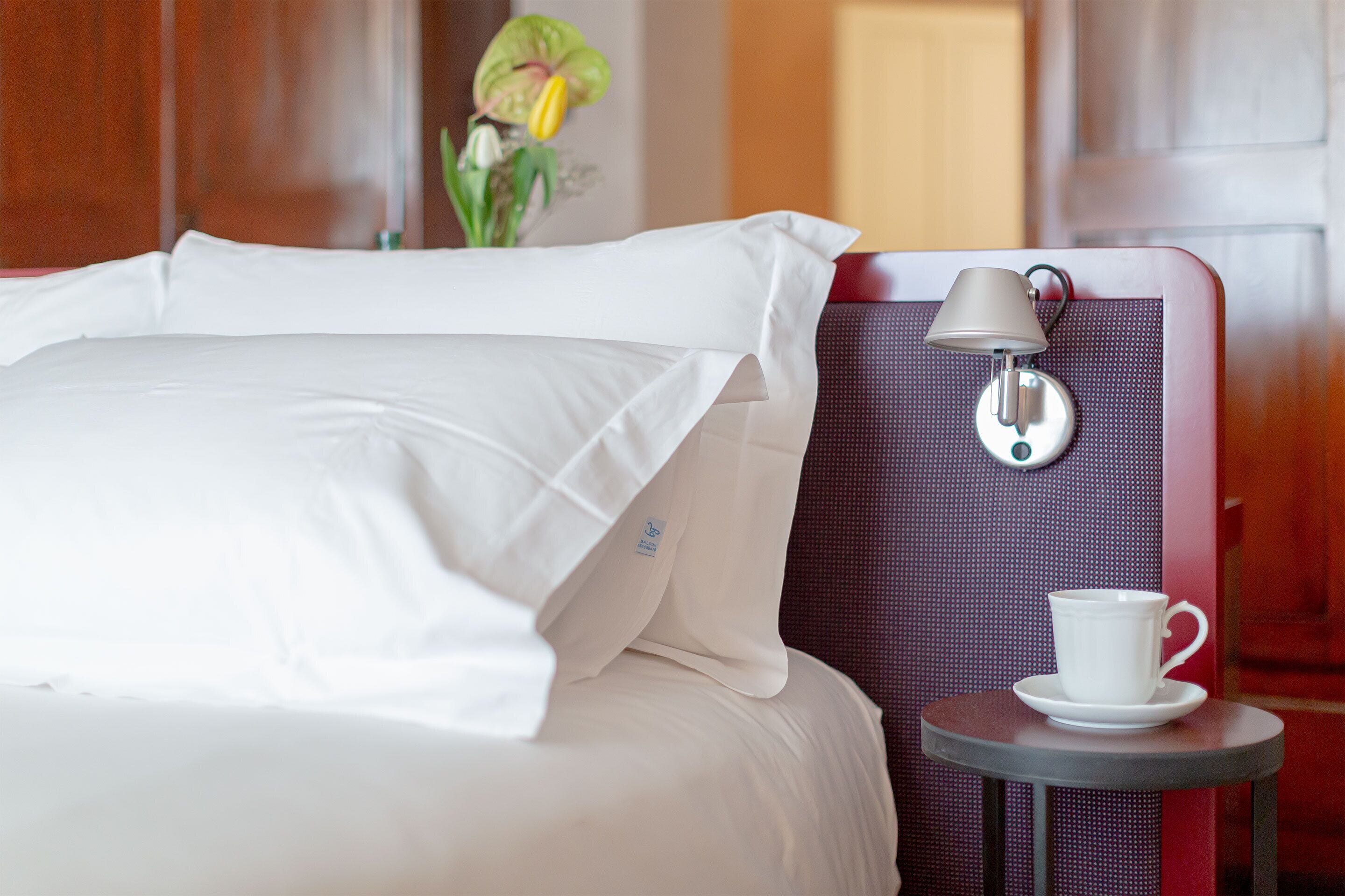 Un lit bien fait, avec du linge de lit blanc, une petite table sur laquelle se trouvent une tasse et une soucoupe blanches, et une lampe fixée au mur.
