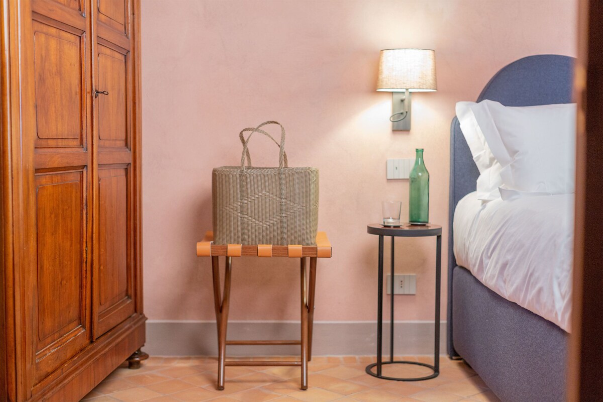 Une armoire en bois, un panier en osier posé sur un tabouret en bois, une petite table avec une lampe et un lit avec du linge de lit blanc.