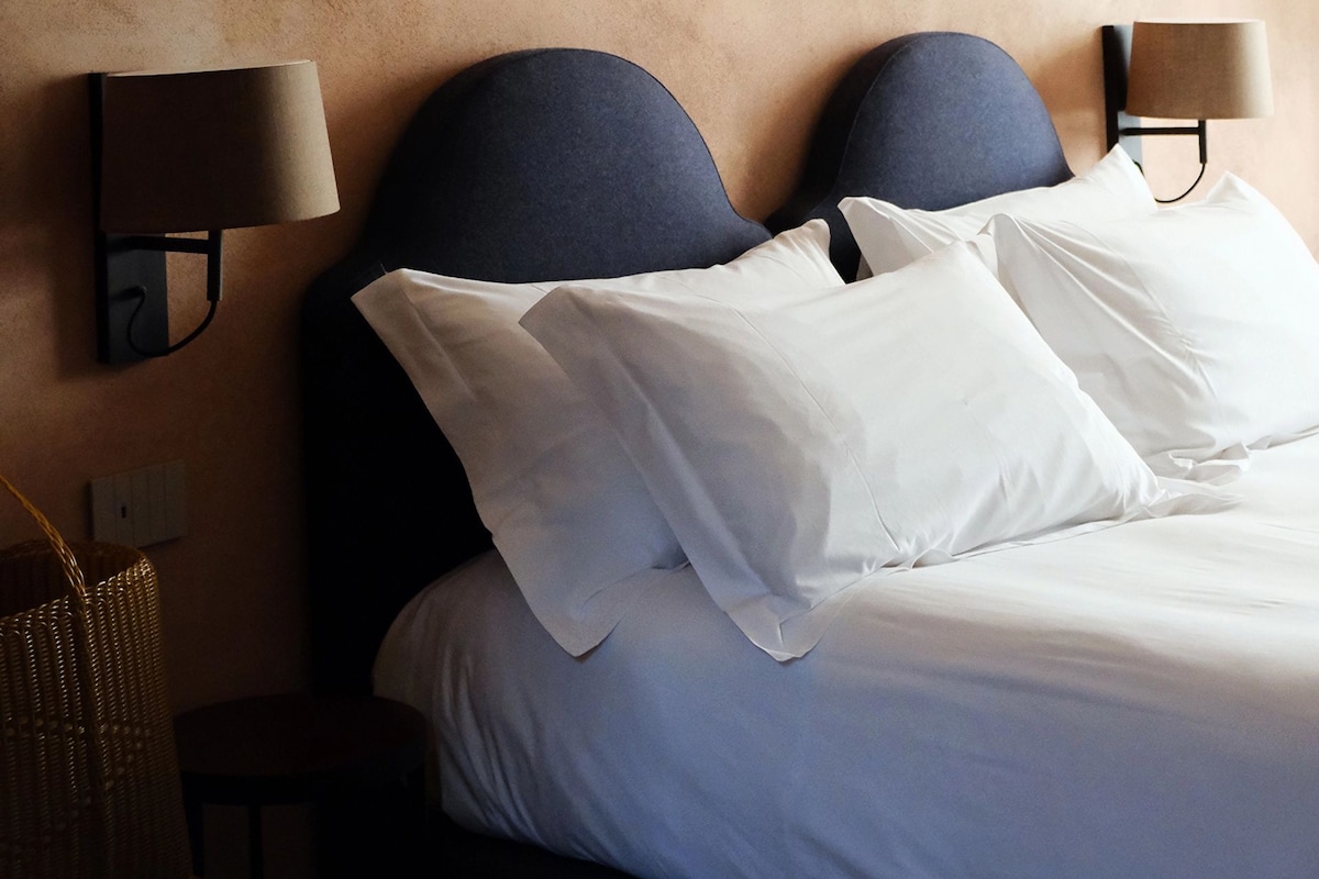 Un lit bien fait, avec du linge de lit blanc et une tête de lit sombre, flanqué de lampes fixées au mur.